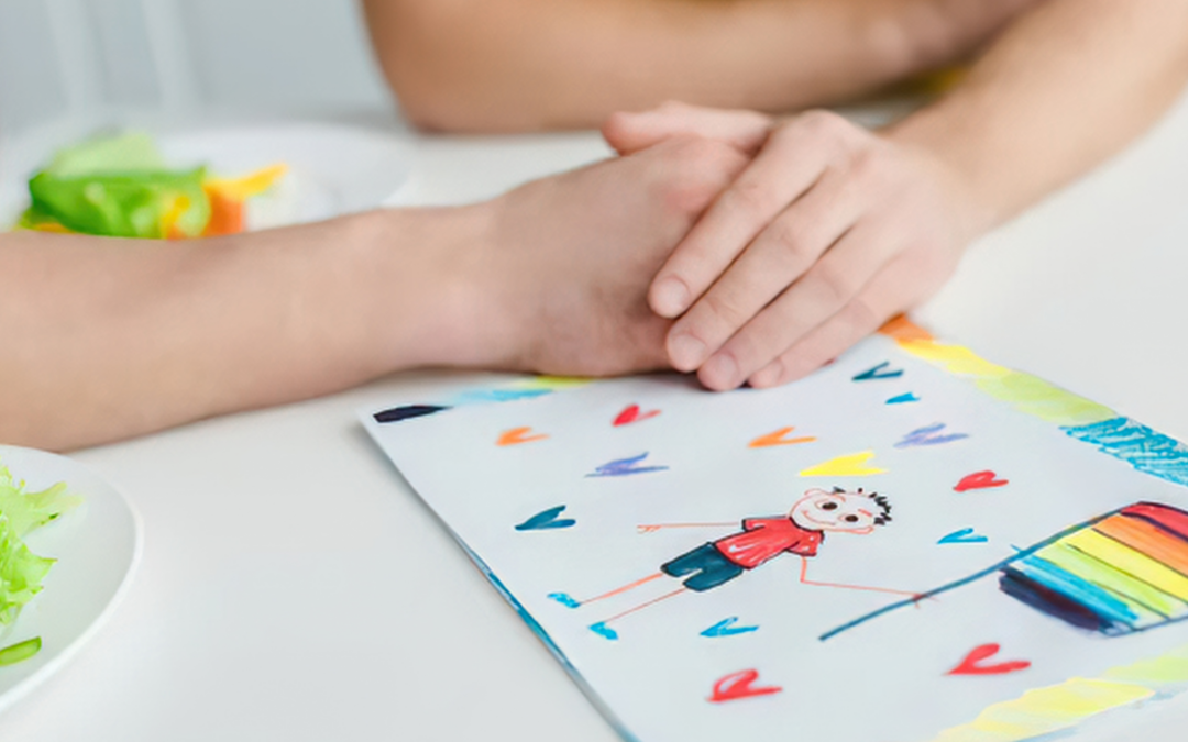A close-up of two hands resting on a table beside a child’s drawing of a stick figure holding a rainbow flag, surrounded by small colorful hearts. A plate with salad sits nearby, creating a warm, supportive scene.